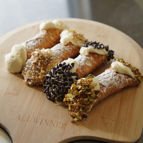 Assortment of cannolis with various toppings like chocolate chips, nuts, and powdered sugar on a wooden board.