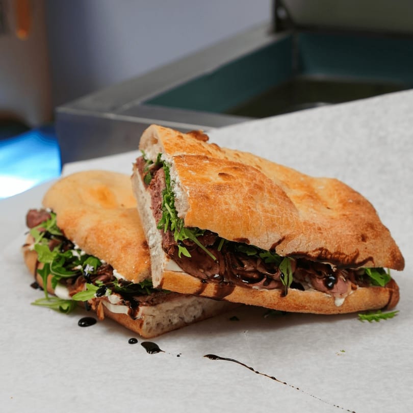 Two halves of a delicious sandwich with meat, arugula, and balsamic glaze on ciabatta bread.