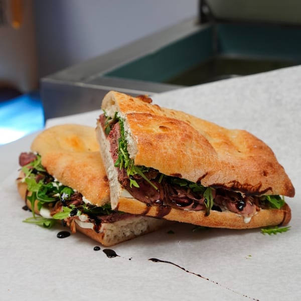 Two halves of a focaccia sandwich filled with roast beef, arugula, cheese, and balsamic glaze.