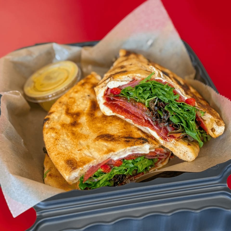 Grilled flatbread sandwich with salami, cheese, arugula, and red peppers, served with a side sauce.