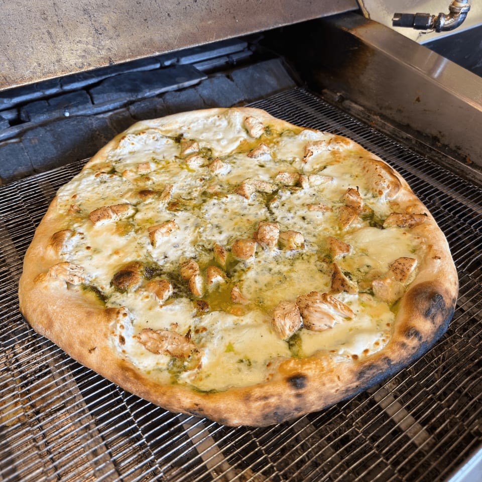 A freshly baked chicken pesto pizza with melted cheese on a metal conveyor belt in a pizza oven.