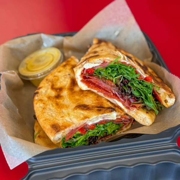 Two halves of a grilled flatbread sandwich filled with meat, cheese, and greens, with a side of sauce.