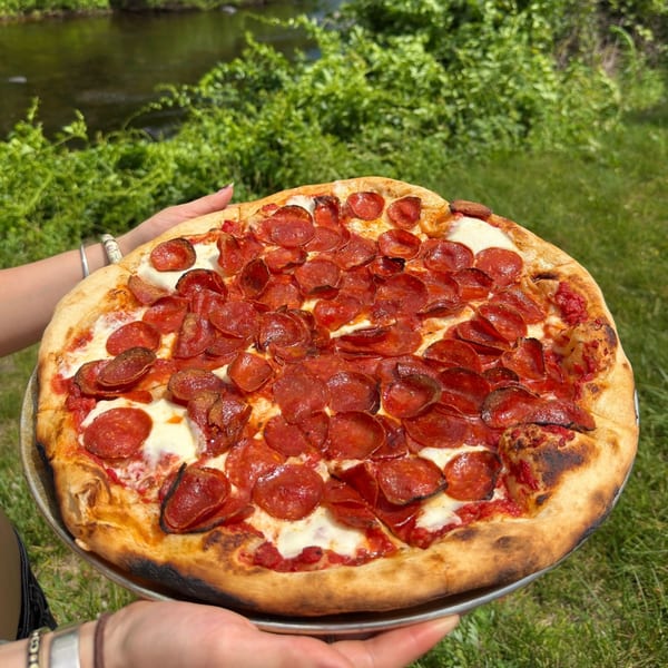 Hands holding a large pepperoni pizza with a bubbly crust, outdoors by a river and green foliage.