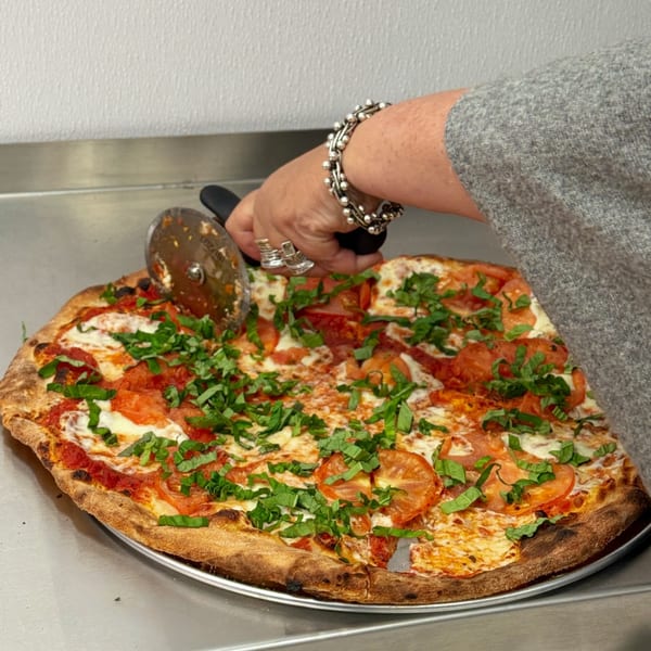 A hand uses a pizza cutter to slice a fresh pizza topped with tomatoes, mozzarella, and basil.