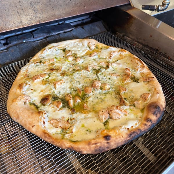 A freshly baked chicken pesto pizza with melted cheese on a metal conveyor belt.