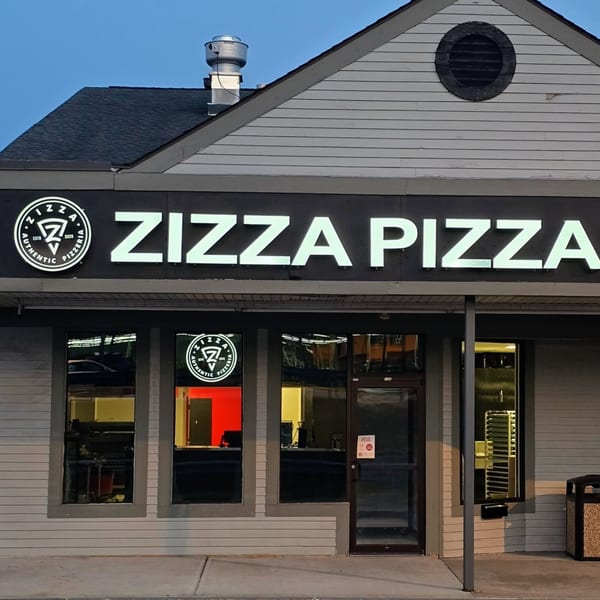 Exterior of Zizza Pizza restaurant at dusk, with glowing white sign and interior lights visible through windows.