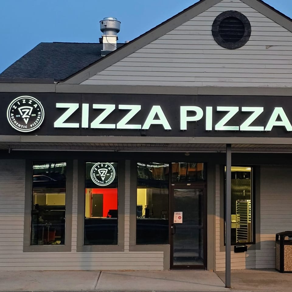 Exterior of Zizza Pizza restaurant at dusk, with glowing white sign and interior lights visible through windows.